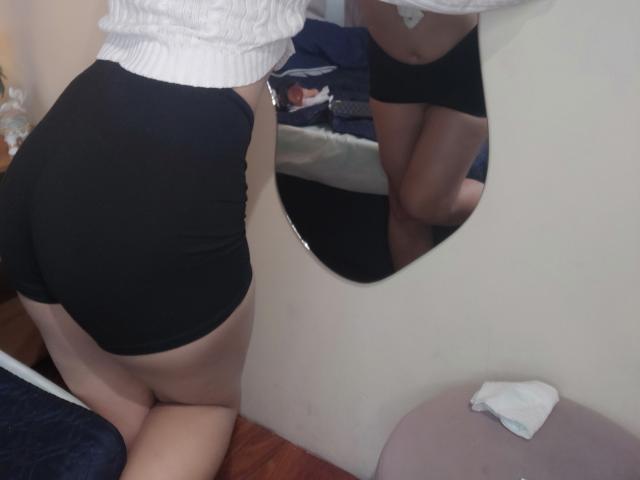 DannyRouds, inginocchiato di fronte a uno specchio, indossa pantaloncini neri attillati e una maglietta bianca, mostrando dei glutei prominenti. riflessi che ne esaltano le curve, un tessuto bianco su una superficie viola.