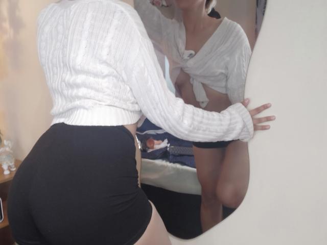 Danny staat voor de spiegel, gekleed in een witte blouse met strik en zwarte shorts, en leunt voorover, waarbij haar decolleté zichtbaar is. reflectie. Hel licht, contrast tussen wit en zwart, sensuele pose, spiegel legt reflectie vast..