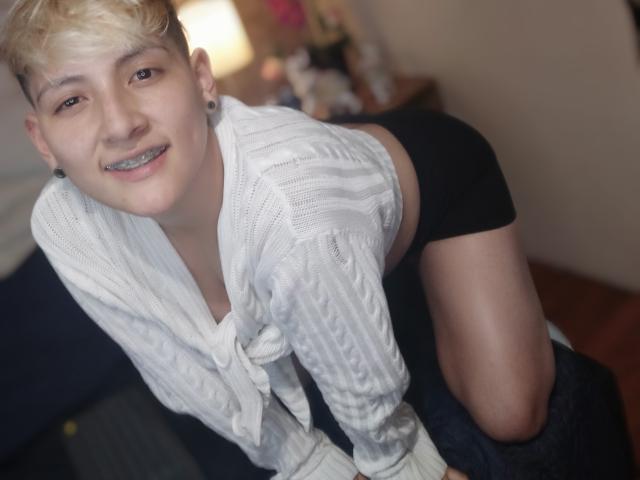 DannyRouds leunt voorover met een grijns, blond haar, een wit gebreid shirt, zwarte shorts, grijze leggings die naar beneden zijn getrokken, waardoor kaalgeschoren dijen. Zacht licht, onscherpe achtergrond, nonchalant, sexy pose.