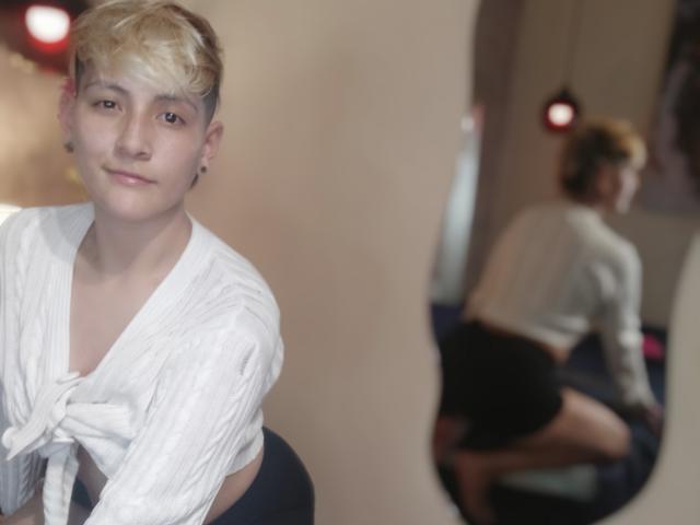 Fotografia di DannyRouds, un giovane con capelli biondi corti, che indossa una camicia bianca annodata e guarda direttamente verso... fotocamera. Il riflesso nello specchio sullo sfondo mostra la schiena di DannyRouds, evidenziando un sedere formoso. Illuminazione soffusa.