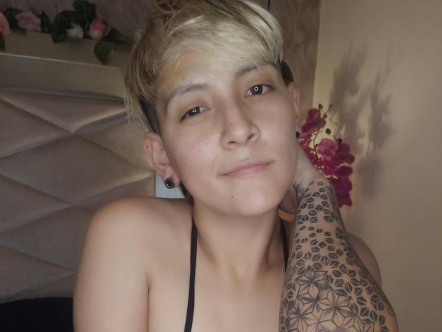 Fotografia de DannyRouds com um corte de cabelo curto e loiro estilo pixie, piercings pretos nas orelhas e uma intrincada tatuagem preta cobrindo o braço. braço direito, vestindo uma blusa preta com alças, olhando diretamente para a câmera com um leve sorriso..