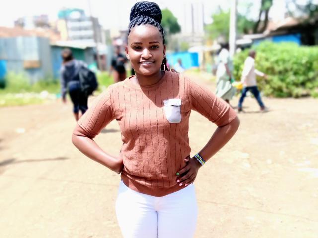 Emphi69 stands in a sunny outdoor setting, hands on hips, wearing a tight, orange, long-sleeve top and white pants, with braided hair, a confident smile, and a watch on her wrist.