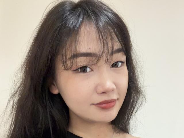 Close-up of PinkyBlooom with long black hair, bangs, and light skin. PinkyBlooom has small breasts, dark eyebrows, and a subtle smile. Background is plain white. Fucking hot, looking straight at camera.