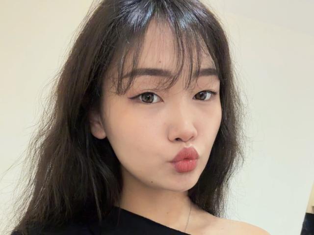 Nahaufnahme von PinkyBlooom mit asiatischen Gesichtszügen, schwarzen Haaren und Pony, die die Lippen zu einem Schmollmund formt und rote Lippen trägt, und die ein schwarzes Oberteil trägt. Helle Haut, leichte Schatten unter den Augen, weißer Hintergrund. Verdammt süß..