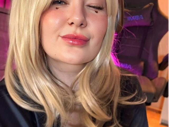 Close-up photo of EffyPamela with blonde hair, glossy pink lips, and a slight smirk. EffyPamela's face has a few small acne marks. Background shows a blurred chair and purple light.
