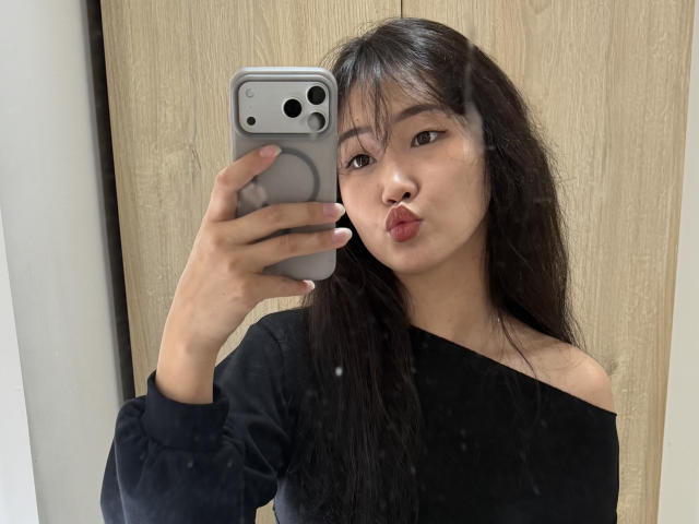 PinkyBlooom, avec ses longs cheveux noirs, prend un selfie, les lèvres pincées, portant un haut noir à une épaule, tenant un iPhone gris. Fond avec texture bois, tension sensuelle, légers reflets de lumière sur le visage..