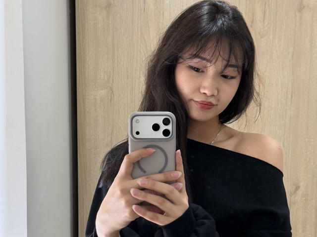 PinkyBlooom, una donna asiatica con lunghi capelli neri, indossa una maglietta nera con una spalla scoperta e tiene in mano un iPhone bianco con una cover grigia. presa (di corrente), che guarda il telefono, sottile tensione sessuale, sfondo neutro.