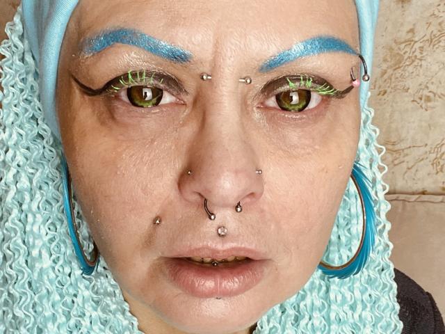 Close-up van het gezicht van Icecandyshokolade, met blauw krullend haar, groene ogen en meerdere piercings in de neus. wenkbrauwen. Grote, blauwe oorringen, serieuze uitdrukking, heldere verlichting..