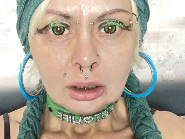 Gros plan du visage d'Icecandyshokolade, montrant plusieurs piercings faciaux : nez, septum et lèvre. Des yeux verts avec du fard à paupières vert, un foulard vert et des boucles d'oreilles cercles bleues. Collier ras du cou vert avec strass.
