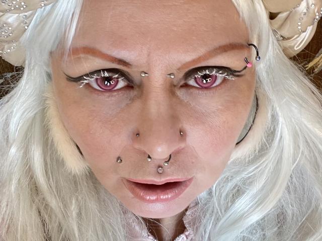 Close-up van het gezicht van Icecandyshokolade, met wit haar, roze ogen en meerdere piercings in het gezicht, waaronder in de neus. ogenbrauwringen. Haar lippen zijn lichtjes open, waardoor een vleugje verleidelijke blik zichtbaar is..