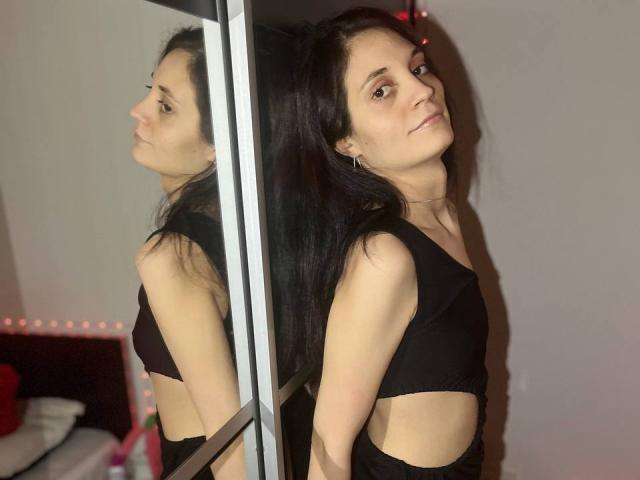 Fotografía de FayeSlate, de pie frente a un espejo, vistiendo una camiseta corta negra, con cabello negro largo, piel pálida y expresión seria. Expresión, reflejo que muestra el perfil lateral, fondo minimalista. Increíblemente atractivo/a..