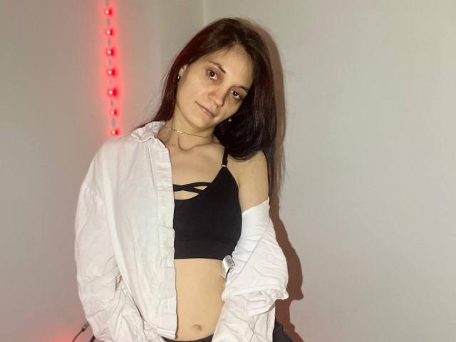 Fotografie von FayeSlate, einer dunkelhaarigen Frau mit heller Haut, die ein offenes weißes Hemd und einen schwarzen BH trägt und an einer weiße Wand mit roten LED-Leuchten. Flirty, verführerische Pose.