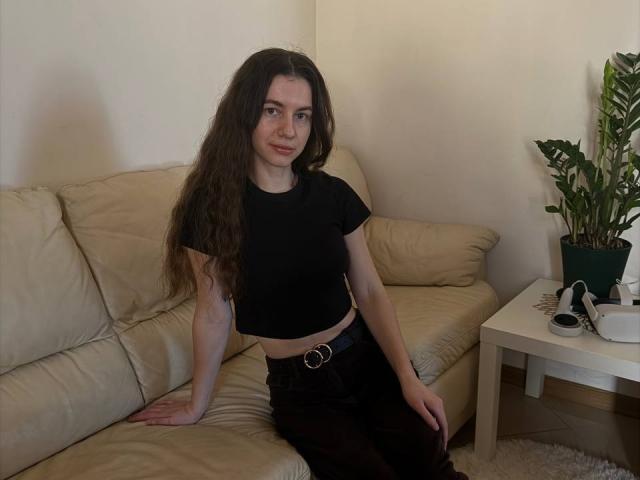 HottyJuli with long, curly brown hair, wearing a black crop top and black skirt, sitting on a beige leather couch. Right arm on cushion, left hand on lap, sexy vibe. Potted plant on white table.
