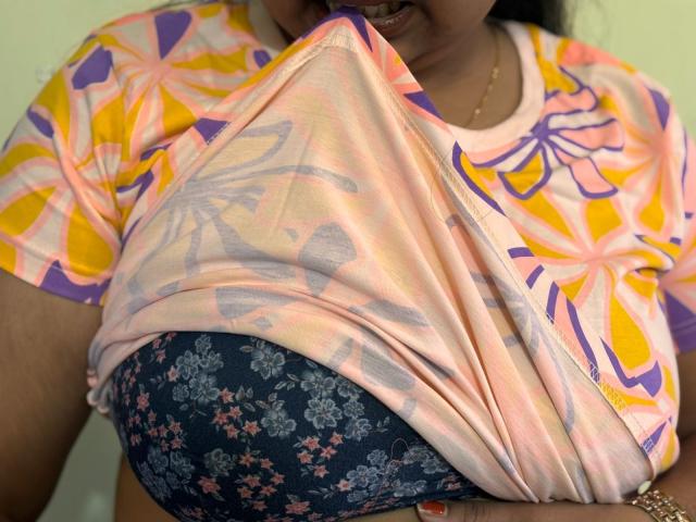 Primer plano de HottyMonat levantando una camisa floral de colores para revelar un sujetador azul oscuro con pequeñas flores rosas. La piel brillante, la tela ajustada y el escote visible crean tensión sexual. Colores brillantes.