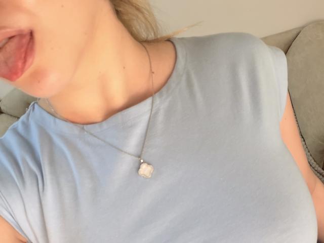 Close-up do tronco superior de SugarrX, mostrando sua camisa azul clara, um colar delicado com um pequeno pingente, e ligeiramente lábios entreabertos, com um leve toque de batom vermelho. Dicas dos dedos visíveis.