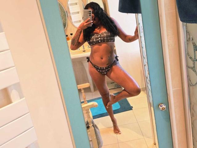 Naatyy prend un selfie devant un miroir, debout avec une jambe levée, et porte un bikini à carreaux noir et blanc. De longs cheveux noirs et ondulés dissimulent en partie son visage. Elle tient un téléphone dans sa main droite..