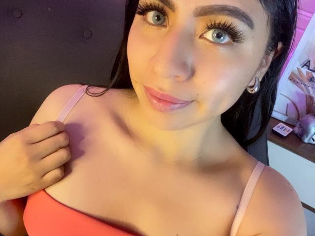 Primo piano di ZoeRedds, una donna dalla pelle chiara con lunghi capelli neri, occhi azzurri e labbra piene, che indossa un top rosa a spalline. Top con spalline. Ha una mano sul petto, sorridendo in modo seducente..