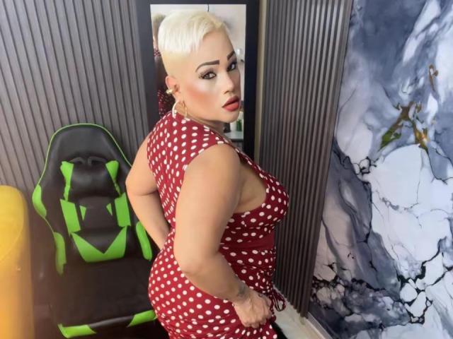 Gianella Cox, bionda con i capelli corti, indossa un vestito rosso a pois e si volta con un'espressione seducente, con un seno generoso. Posizione seducente, appoggiata a un muro crepato, sedia nera con cinghie verdi..