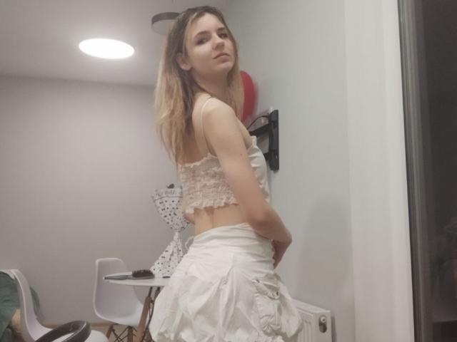 LovieSchryer, debout, le dos à la caméra, porte un soutien-gorge blanc en dentelle et une jupe blanche brillante, et regarde par-dessus son épaule, une main. à la mode. Cheveux blonds, pose décontractée, pièce avec des meubles modernes..