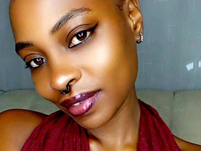 Close-up de Queen-Adora, uma mulher atraente, de pele escura e lábios roxos brilhantes, com delineador preto, um pequeno piercing no nariz e brincos. Ela olha de forma sedutora, com um leve inclinar da cabeça, vestindo uma blusa vermelha de alças..