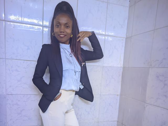 Victorine Stone se tient devant un mur carrelé, vêtue d'un blazer noir par-dessus une chemise blanche et un pantalon blanc. Elle a la peau foncée, une queue de cheval haute et un sourire confiant. Une main dans la poche, le bras levé..