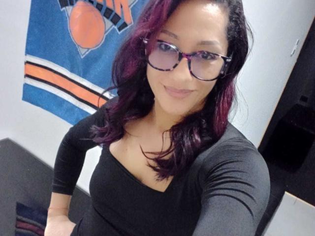 Photograph of SamanthaCorine, wearing black glasses and a tight black top, with shoulder-length dark red hair, sitting against a wall with a sports flag. Cock-sized cleavage, sultry smile.