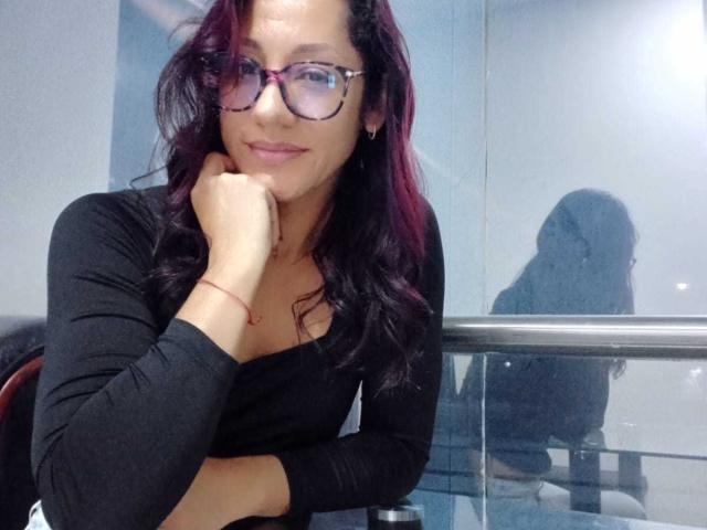 Photograph of SamanthaCorine with long dark hair, wearing black glasses and a tight black top, leaning on her hand with a slight smile, reflective glass behind her, shadow on the glass.