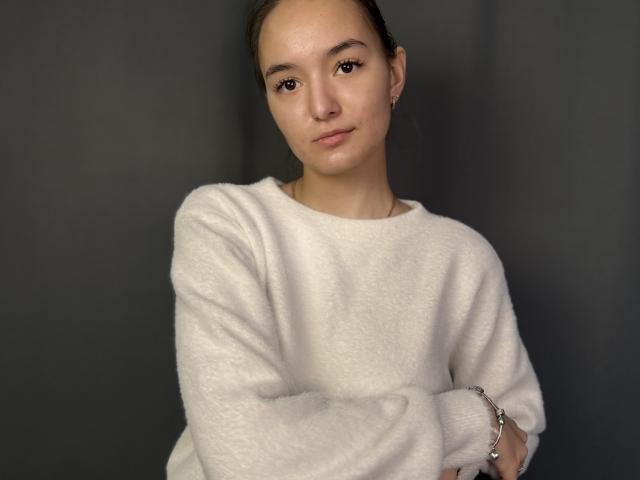 Photograph of LaritaDeralph with a light skin tone, wearing a white, fluffy sweater, arms crossed, looking directly at the camera with a neutral expression, dark background, slight sexual tension.