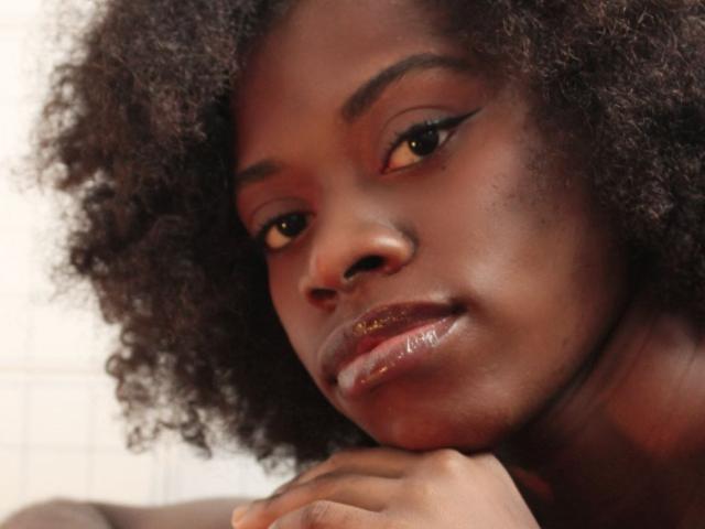 Gros plan de Vsakurap, une femme à la peau foncée, avec des cheveux noirs bouclés, des lèvres brunes brillantes et un trait d'eye-liner ailé. Elle pose son menton sur sa main, regardant directement l'appareil photo avec une expression sexy et aguicheuse..
