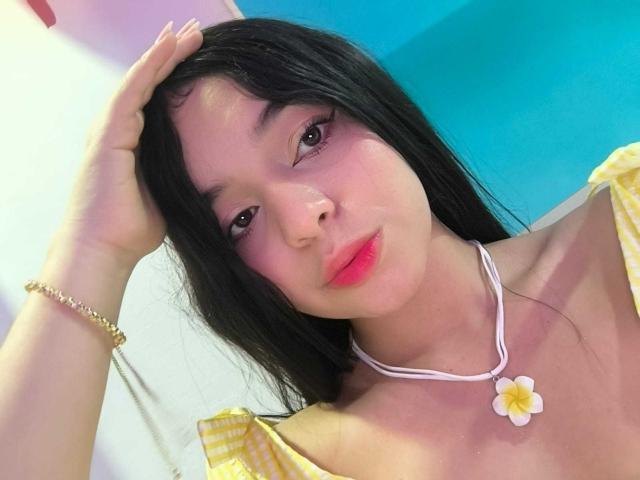 Lissy Carter tem cabelo preto, pele clara e lábios vermelhos. Ela está vestindo uma blusa amarela com um ombro de fora e um colar branco com uma margarida. A mão dela está no cabelo, demonstrando tensão..