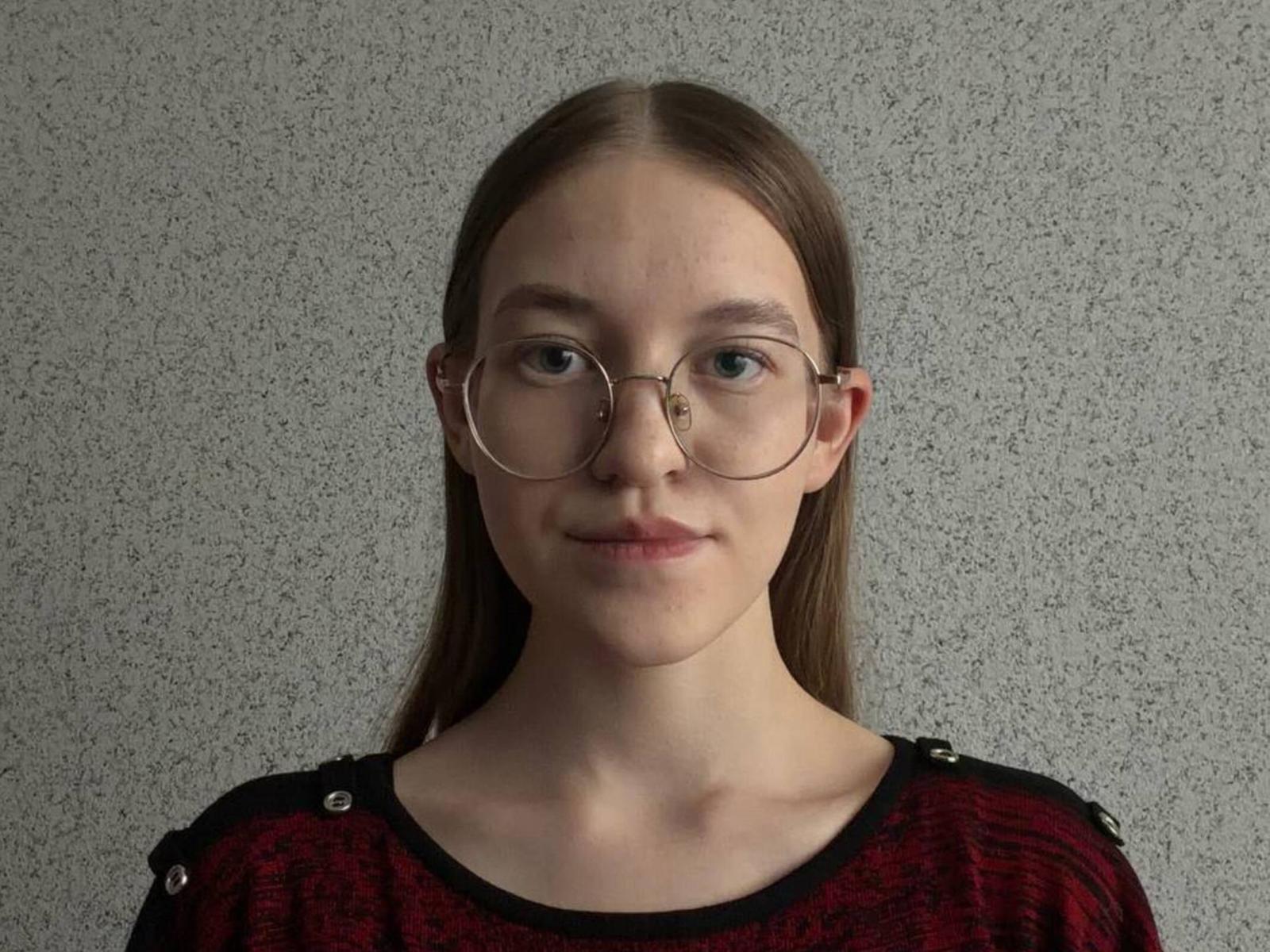 Photographie de Toccara Pacholec, avec de longs cheveux bruns, portant des lunettes rondes et un haut rouge à motifs, regardant la caméra. Avec un léger sourire, un éclairage doux, un fond gris texturé. Tellement mignon..