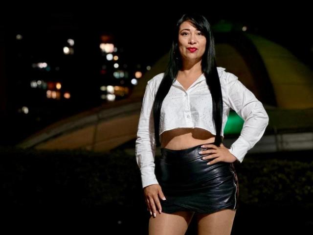 Karimelee stands confidently in a night scene, wearing a white button-up crop top and tight black leather miniskirt. Her long black hair is straight, and she has red lipstick. She's posing with one hand on her hip, showing off her toned midriff. City lights in the background.