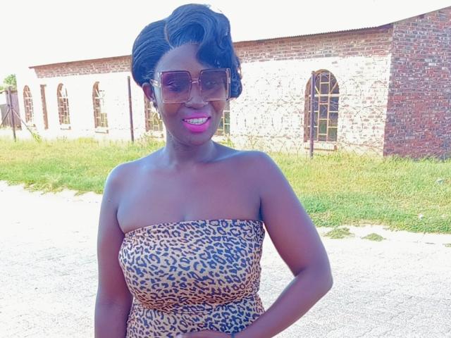 SmokinPhatMellons with dark skin and short, curly black hair, wearing a strapless leopard-print dress and large sunglasses, smiling brightly. Brick building with arched windows in the background. Sunlit, vibrant.