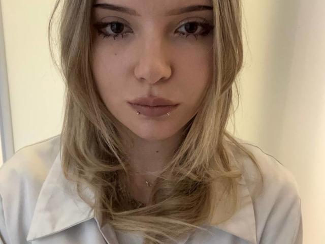 Close-up van ElidiaKrotine, een blonde vrouw met blauwe ogen, die een witte blouse draagt. Ze heeft een lip-piercing en een subtiele, intense uitdrukking. Zacht licht accentueert haar gezichtsvorm..
