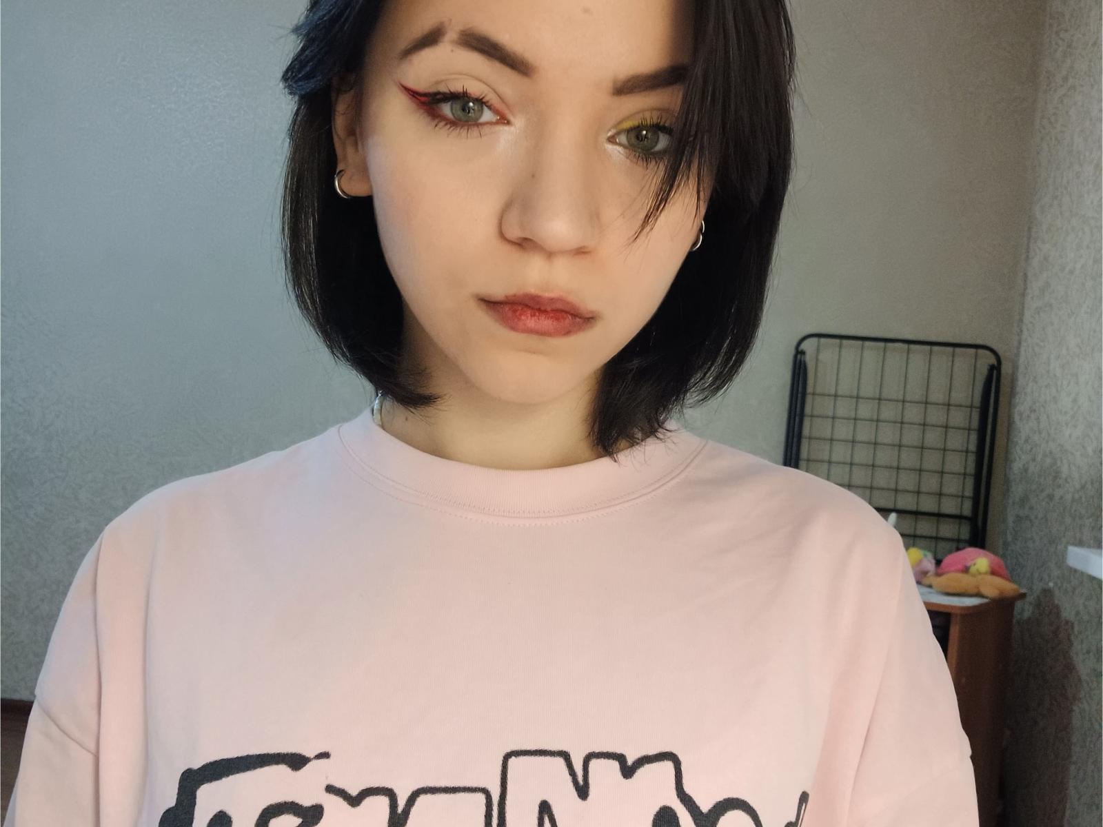 HottieFlemmings tem cabelo preto curto, olhos verdes com delineador marcante e batom vermelho. Usando um moletom branco com uma estampa preta. Uma luz suave realça sua pele clara. Olhar muito sério..