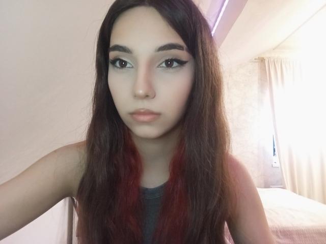 Photographie de Tisha Kreisman avec de longs cheveux bruns foncés, un trait d'eye-liner noir épais et une peau pâle. Elle regarde directement l'appareil photo avec une expression neutre, et elle porte un haut gris foncé. Fond clair.