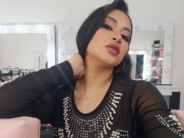 Adalina Vera, avec ses cheveux noirs, son rouge à lèvres foncé et son maquillage prononcé, porte un haut noir orné de perles blanches, se penchant sur un bras, regardant de manière séduisante l'appareil photo, portant un collier en or..