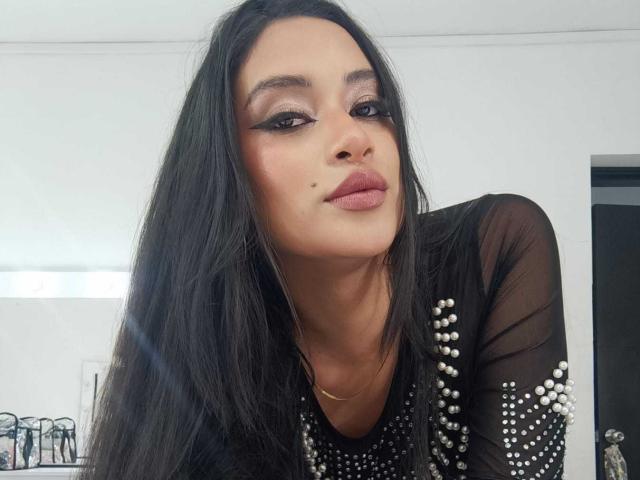 Close-up van AdalinaVera, een vrouw met lang zwart haar, die een zwarte doorschijnende top draagt met zilveren studs en recht in de camera kijkt. de camera met verleidelijke, halfgesloten ogen, en roze lippenstift.