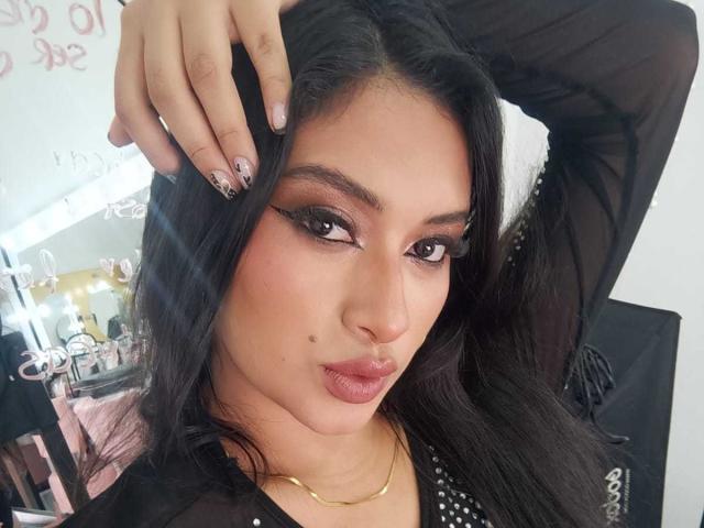 Autoritratto ravvicinato di AdalinaVera, donna dai capelli scuri con un trucco occhi marcato, labbra piene, una lentiggine sulla guancia sinistra, mano nera. capelli, top trasparente nero, collana dorata, espressione sicura, illuminazione intensa.