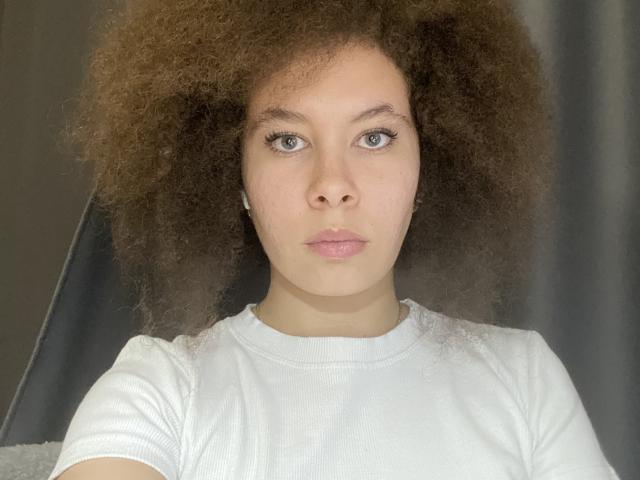 Close-up photograph of ClotildeMerzlak, a young woman with light brown skin and voluminous curly brown hair. She wears a white ribbed shirt, looking directly at the camera with a neutral expression. Fucking hot, with intense blue eyes and slightly parted pink lips.