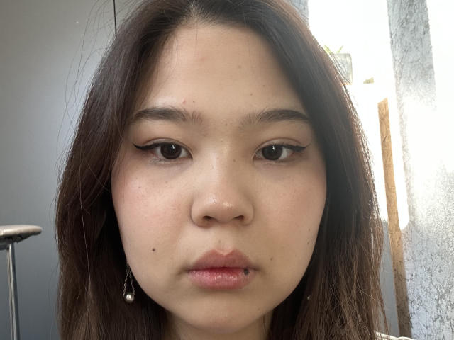 Close-up photo of ShericeKisler with light skin, dark brown hair, and black eyeliner. She has a mole on her cheek, small earrings, and slightly parted lips. Bright light behind her.