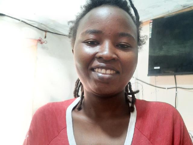 Close-up of RachieChristina, dark-skinned woman with braided hair, smiling, wearing a red shirt with white collar. Bright lighting, neutral background, slight sheen on her face, relaxed expression.