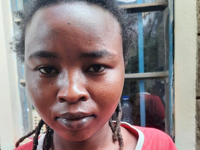 Photographie en gros plan de RachieChristina, avec une peau brun foncé, un visage brillant et de courtes dreadlocks. Elle porte une chemise rouge, et regarde directement l'appareil photo avec une bouche légèrement ouverte, laissant entrevoir quelques dents. L'arrière-plan comprend une fenêtre..