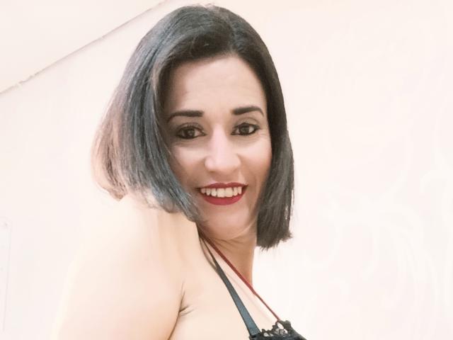 Fotografia em close-up de MaraRoberttss, uma mulher sorridente com cabelo preto curto, batom vermelho e olhos escuros, vestindo uma blusa preta de renda, olhando para a câmera com uma expressão sedutora..