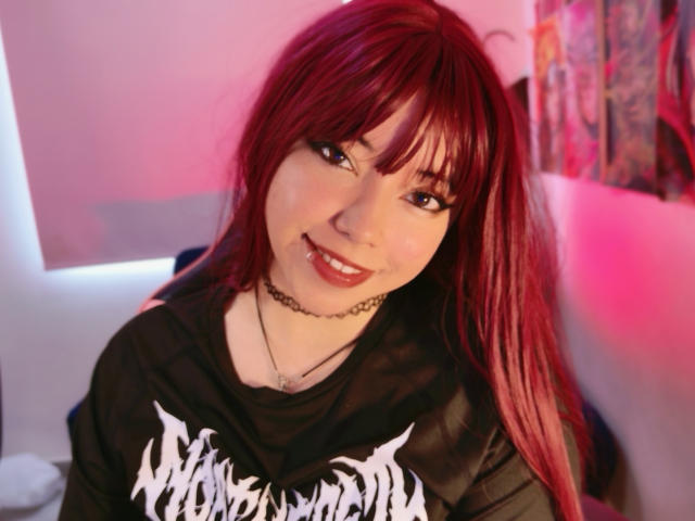 Fotografía de Arieyaara con cabello rojo, flequillo y una camisa negra con texto gráfico blanco. Está sonriendo, lleva un collar y mira directamente a la cámara con una evidente tensión sexual. Iluminación en tonos rosa y rojo.