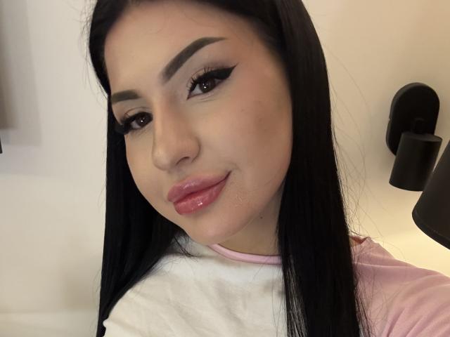 Primo piano di AuraXo, una donna latina con lunghi capelli neri, un pesante trucco occhi nero e labbra rosa lucide. Indossa una maglietta bianca e rosa, e guarda la fotocamera con un leggero sorriso. La sua pelle è liscia, e sullo sfondo si vede una parete nera montata a muro. Molto attraente..