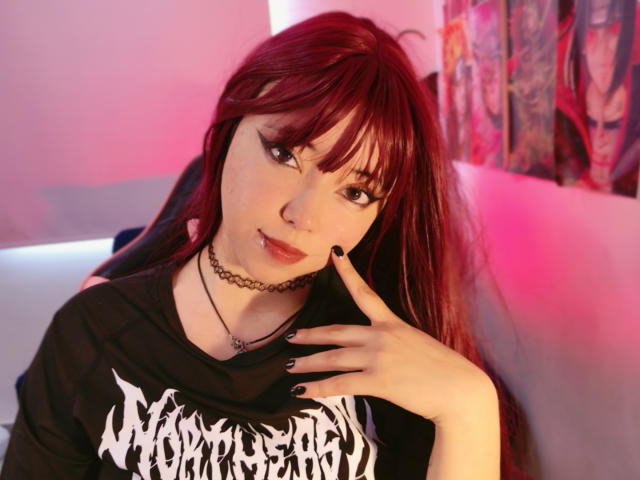 Arieyaara, with red hair and black bangs, wears a black "Slayer" shirt, choker necklace, and black nail polish. She's touching her chin, lips slightly parted, in pink-lit room. Fucking sexy.