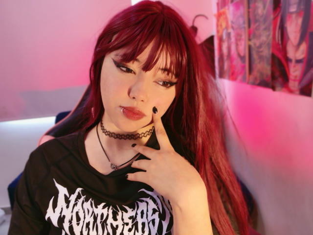 Fotografie von Arieyaara mit langen, roten Haaren, einem schwarzen Choker und einem schwarzen "Metallica"-T-Shirt, die sich an ihrem Schlüsselbein und ihrer Lippe festhält. durchdringender, verführerischer Blick, rosa und rote Beleuchtung, tätowierter Arm im Hintergrund. Verdammt heiß..