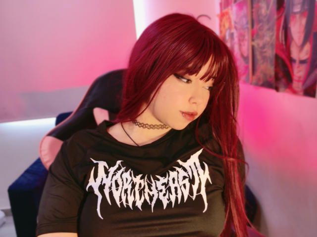 Arieyaara, uma mulher de pele clara com cabelos longos e ruivos, com franja, veste uma camiseta preta da banda "Avenged Sevenfold" e um colar. Inclinando-se para frente com um olhar ligeiramente sedutor, o fundo desfocado com luz rosa..