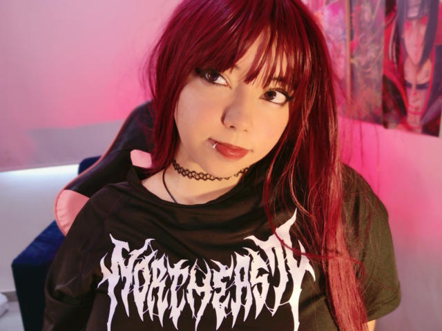 Arieyaara, con piel pálida, cabello rojo y flequillo negro, vestida con una camiseta negra de "Kreator" y un collar, mirando de lado. Una leve sonrisa, iluminación rosa y roja en el fondo..